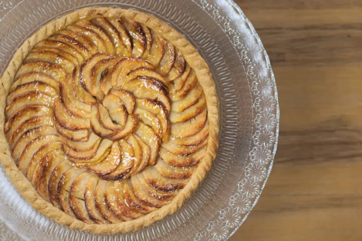 Une magnifique tarte aux pommes, aux pommes caramélisées disposées en spirale sur une pâte dorée, servie sur un plat en verre décoré.