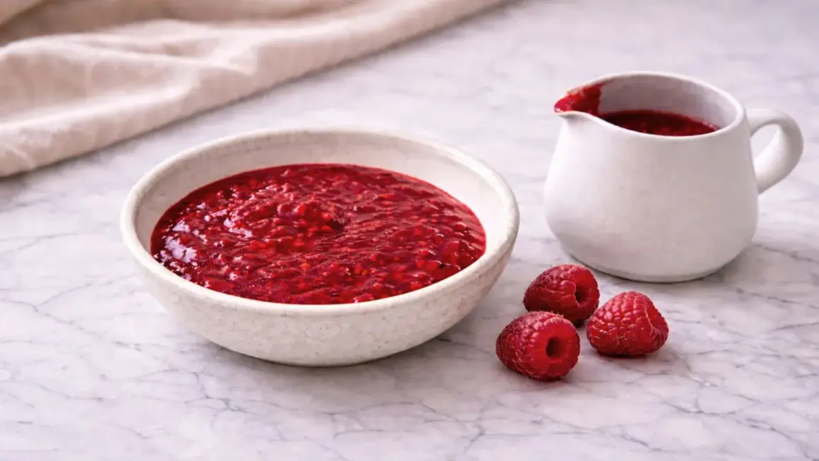 Un bol et un pichet remplis de coulis de framboise, avec trois framboises fraîches à côté.
