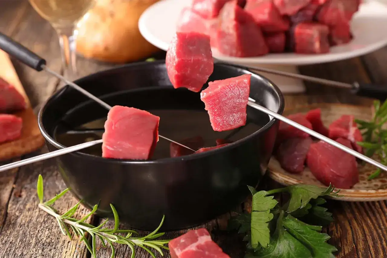 Des cubes de b&oelig;uf sur des brochettes trempent dans l'huile chaude pour une fondue bourguignonne. Un verre de vin blanc et des herbes compl&egrave;tent la sc&egrave;ne.