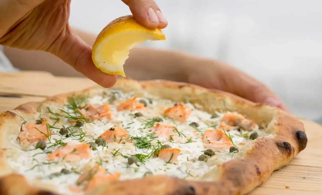 Une main presse un quartier de citron sur une pizza saumon garnie de c&acirc;pres et d'aneth.