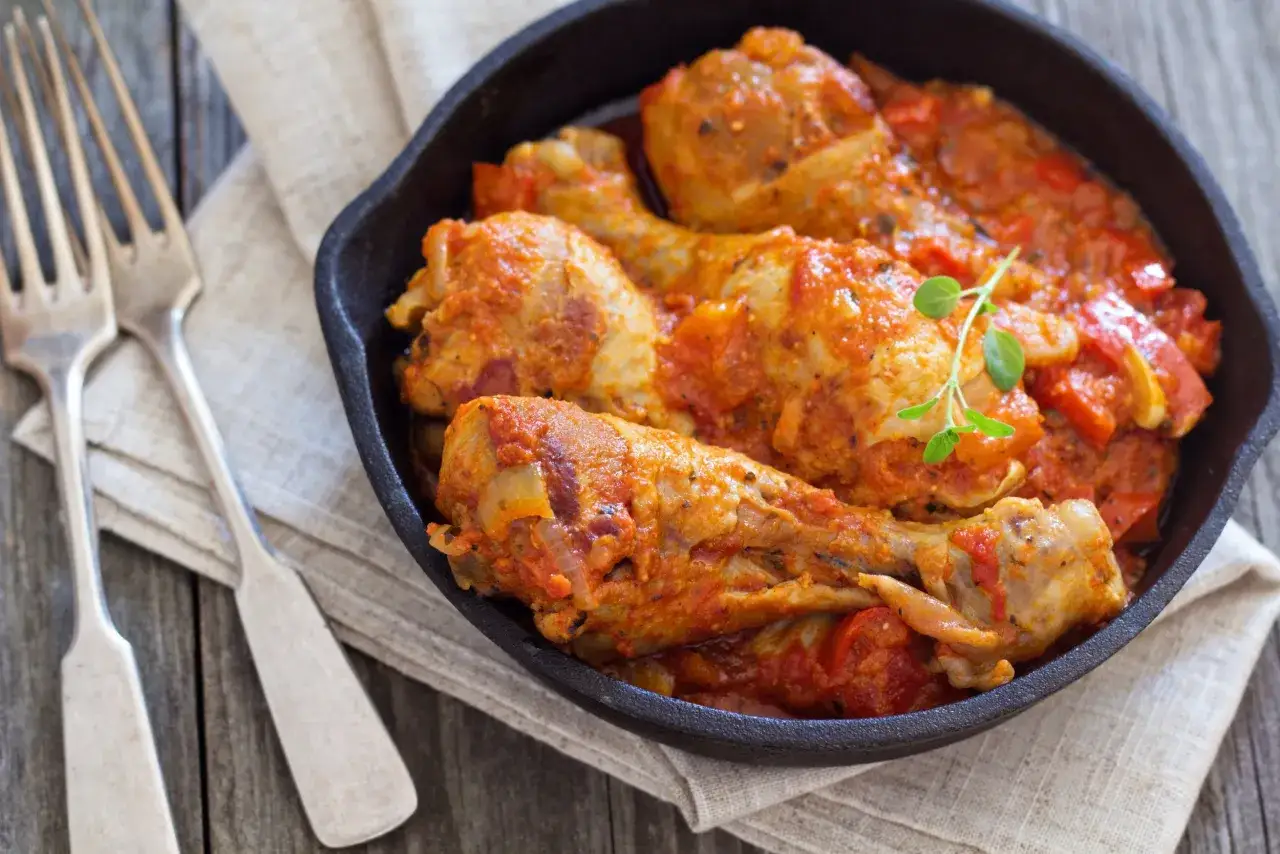Un plat de poulet basquaise mijoté dans une sauce tomate et poivrons, servi dans une poêle en fonte, avec des fourchettes à côté.
