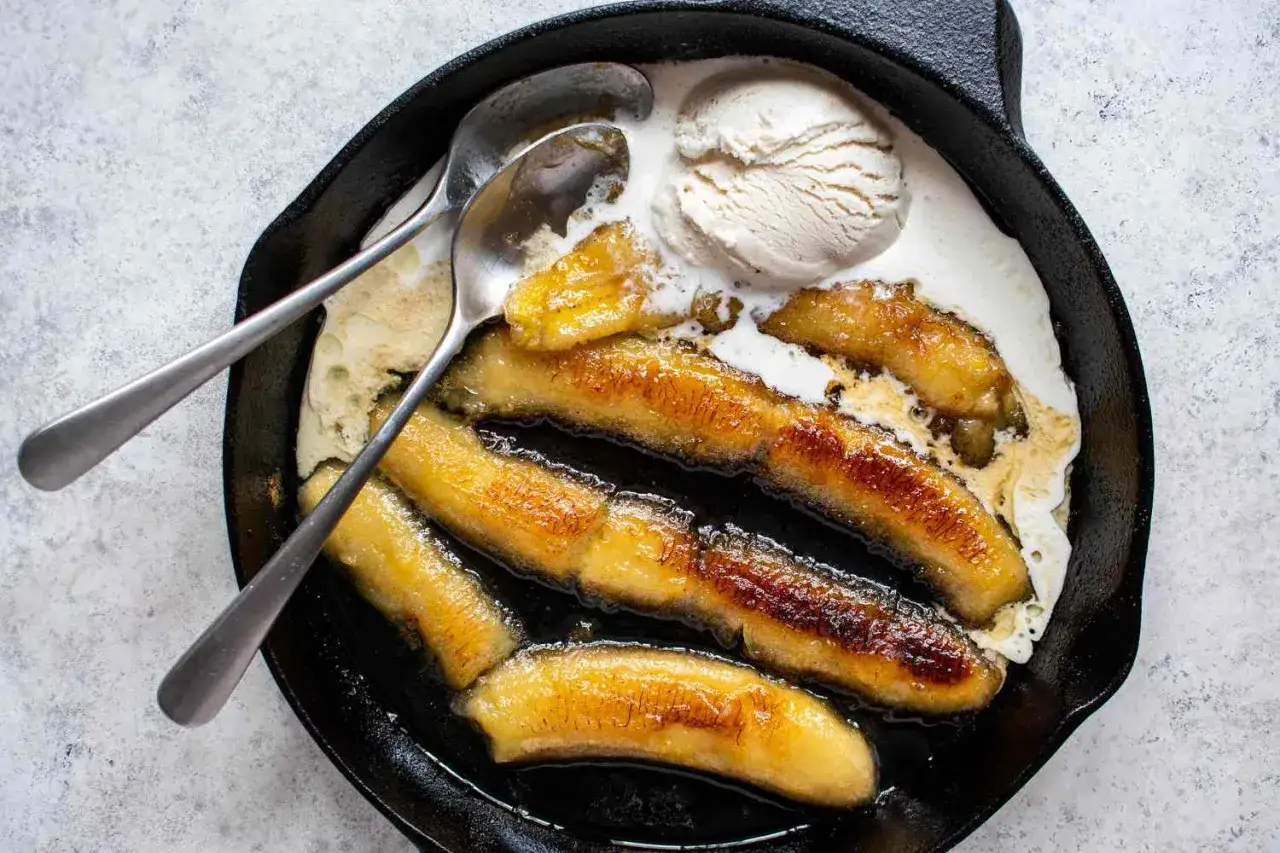 Bananes flambées caramélisées dans une poêle en fonte, servies avec une boule de glace à la vanille et deux cuillères.