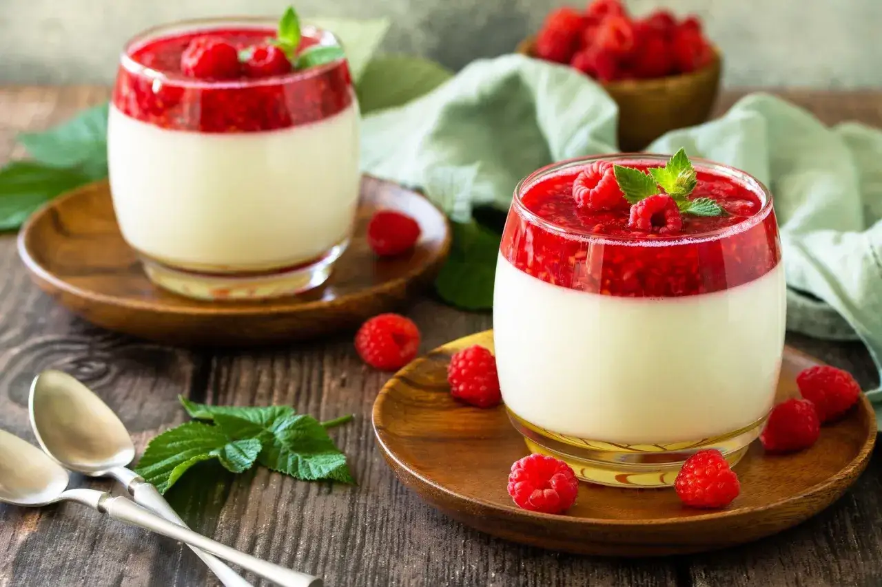 Deux panna cotta crémeuses, surmontées d'un coulis de framboises et décorées de feuilles de menthe. Des framboises fraîches parsèment la table en bois.