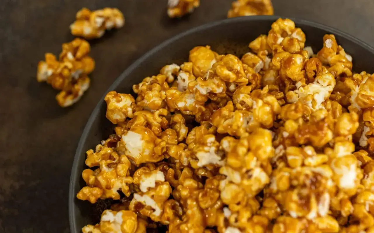 Un bol rempli de pop corn sucré, enrobé d'une sauce caramel dorée et brillante. Quelques grains s'échappent sur le fond sombre.