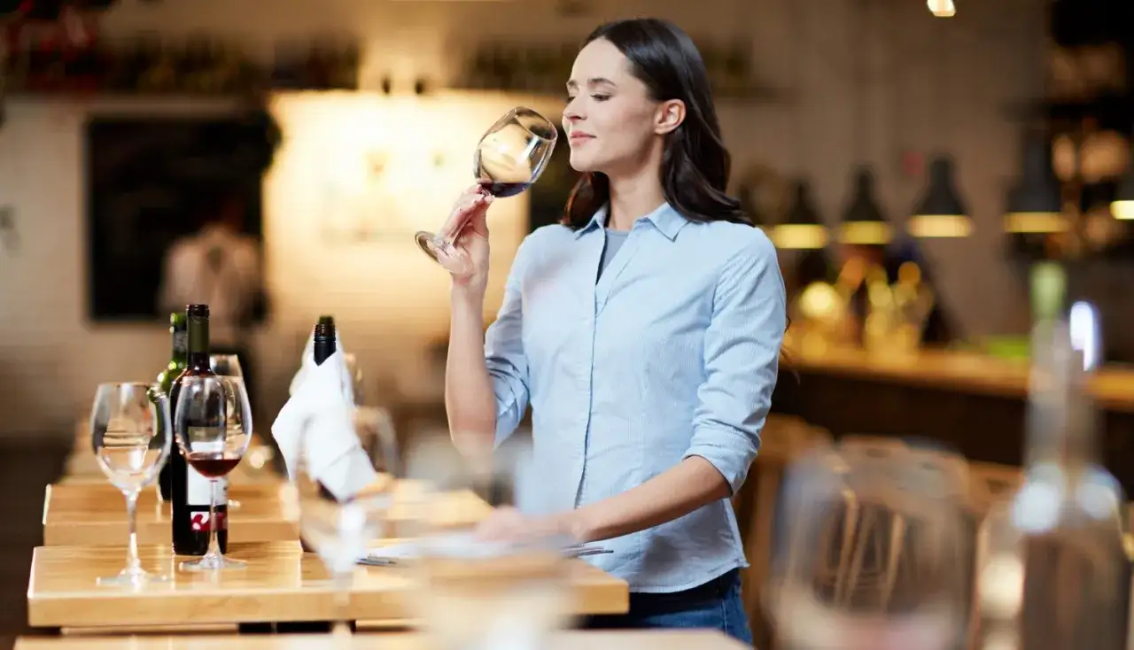 Une femme hume un verre de vin rouge, une &eacute;tape cl&eacute; pour savoir si un vin est bon. Des bouteilles et verres sont dispos&eacute;s sur une table.