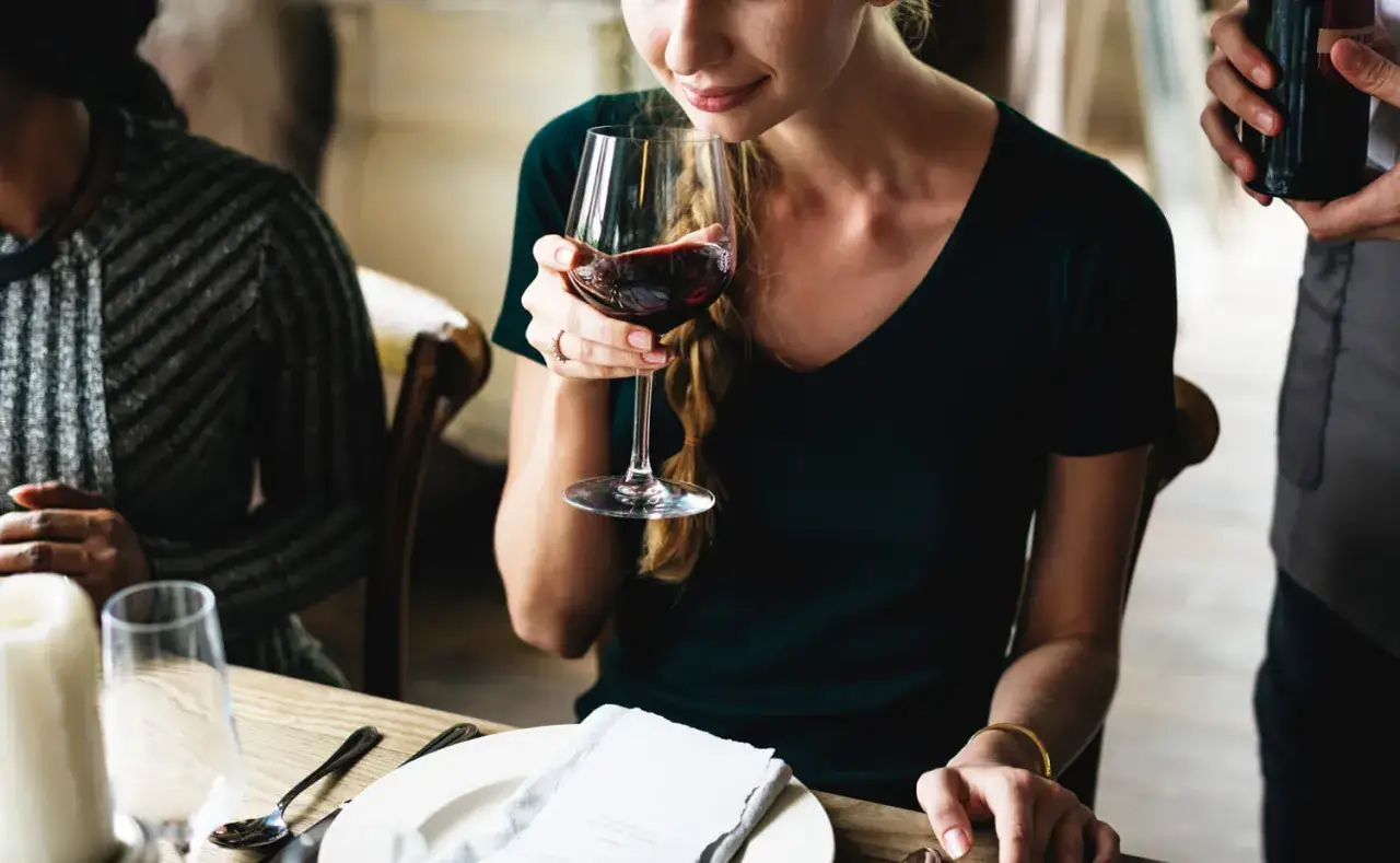 Une femme hume un verre de vin rouge, espérant qu'il ne soit pas un vin blanc bouchonné. Une bouteille est tenue par un serveur.