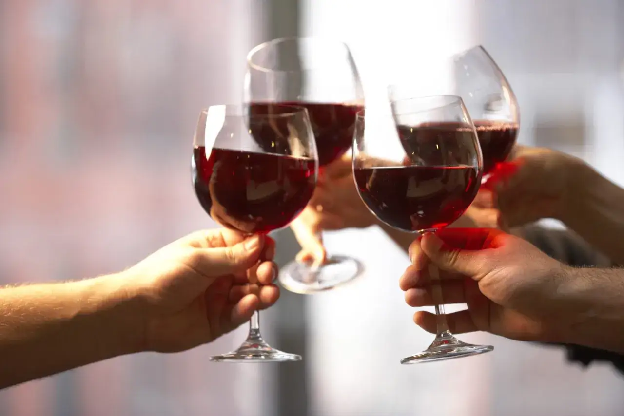 Quatre mains portent des verres de vin rouge, leur couleur profonde capturant la lumi&egrave;re. Un toast &agrave; la joie.