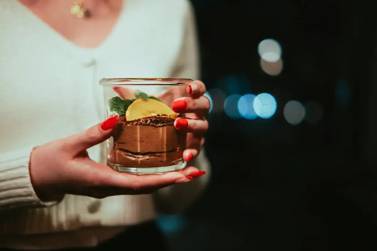 Mains délicatement posées sur un verre de mousse au chocolat, garnie d'une tranche d'orange et de menthe.