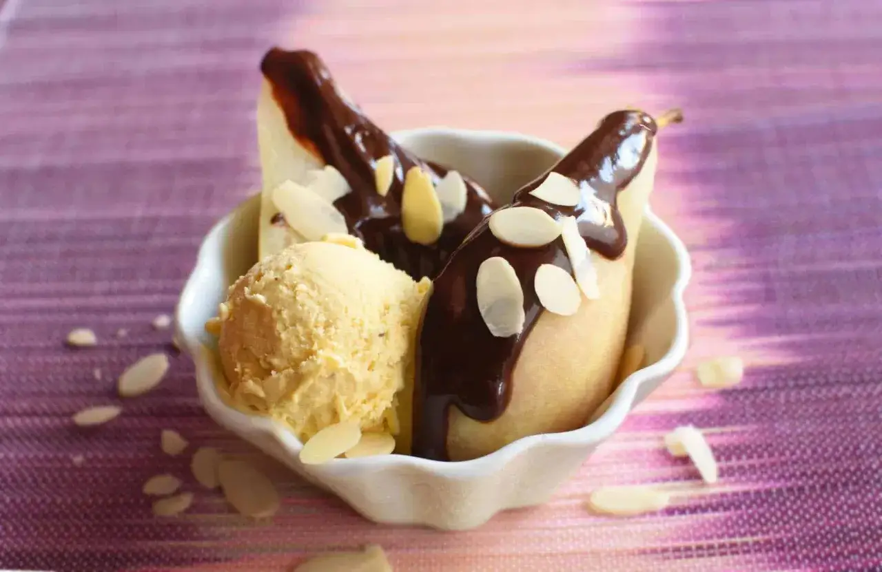 Une poire Belle H&eacute;l&egrave;ne, napp&eacute;e de chocolat chaud et garnie d'amandes effil&eacute;es, servie avec une boule de glace &agrave; la vanille dans un bol blanc.