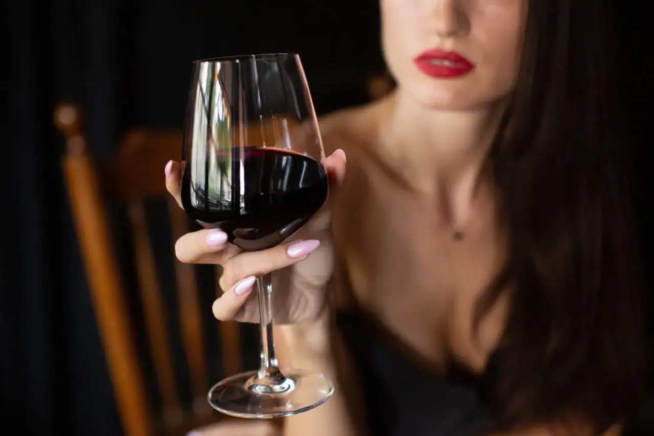 Une main aux ongles vernis tient un verre de vin rouge, montrant comment tenir un verre de vin par la tige pour apprécier la couleur et les arômes.