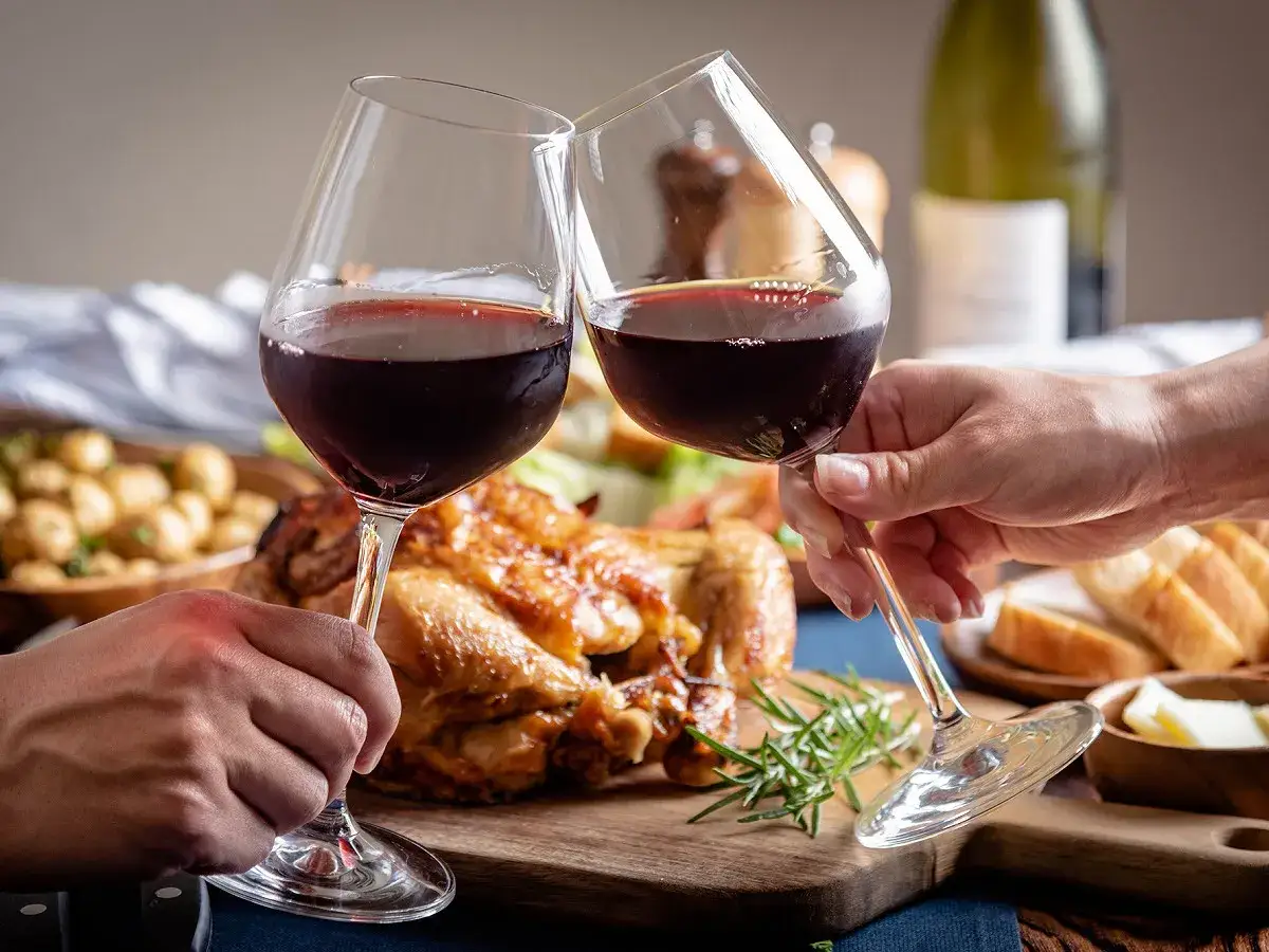 Deux verres de vin rouge levés en toast devant une dinde rôtie, accompagnés de pommes de terre et de pain. Un repas festif.
