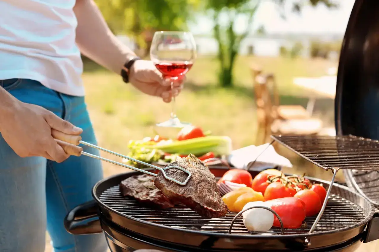 Un homme prépare un barbecue avec des steaks et des légumes, un verre de vin rouge à la main.