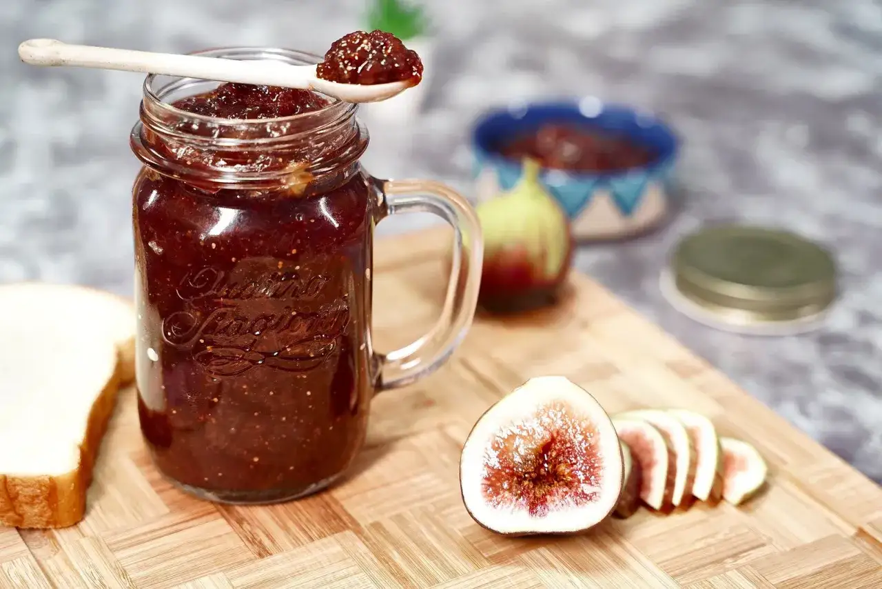 Un pot de confiture de figues maison, une cuill&egrave;re garnie, des figues fra&icirc;ches coup&eacute;es et une tranche de pain sur une planche en bois.