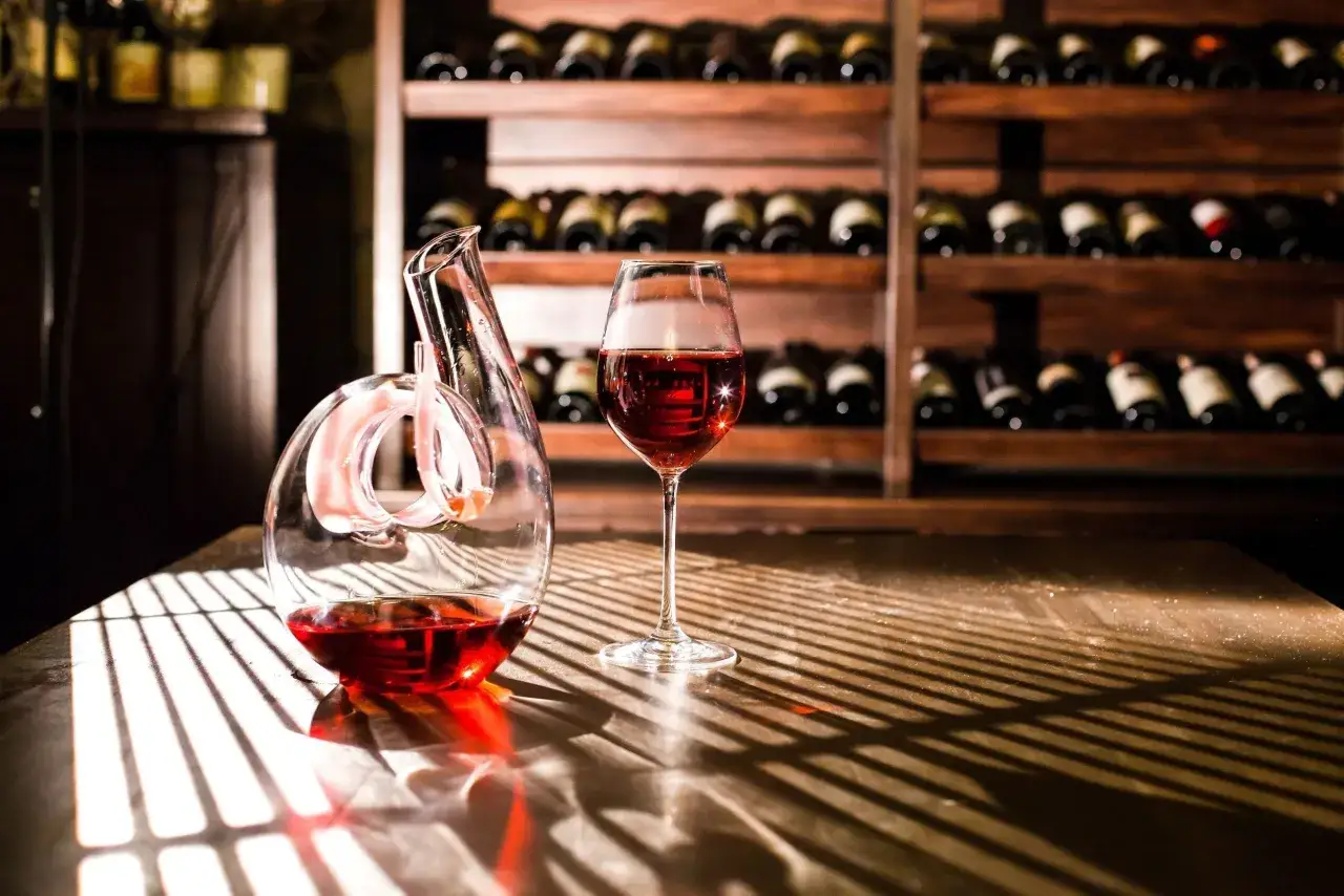 Carafe et verre de vin rouge sur une table, avec des &eacute;tag&egrave;res de bouteilles en arri&egrave;re-plan. La lumi&egrave;re cr&eacute;e des ombres ray&eacute;es, rappelant la lie de vin.