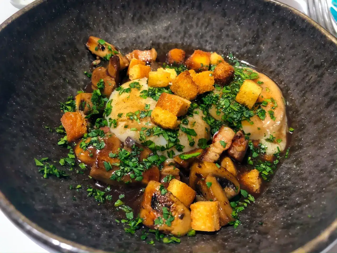 Un œuf meurette, poché et nappé d'une sauce riche, garni de champignons sautés, de lardons croustillants et de croûtons dorés, le tout saupoudré de persil frais.