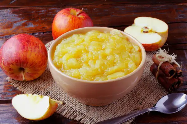 Un bol de compote de pommes maison, entouré de pommes fraîches et de bâtons de cannelle, sur une table en bois rustique.