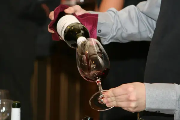 Un serveur verse du vin rouge dans un verre. Le **dépôt de vin blanc** sur la bouteille est visible.
