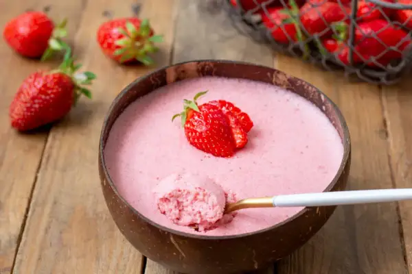 Délicieuse mousse fraise dans une noix de coco, garnie d'une fraise fraîche. Une cuillère dorée en extrait une bouchée aérienne.
