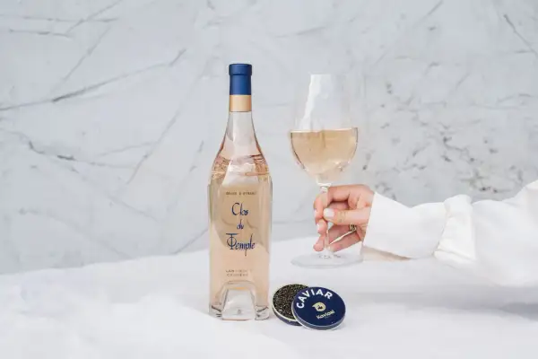 Une main tient un verre de vin rosé, à côté d'une bouteille et de caviar. Le danger du vin rosé est qu'il est si délicieux qu'on en redemande.