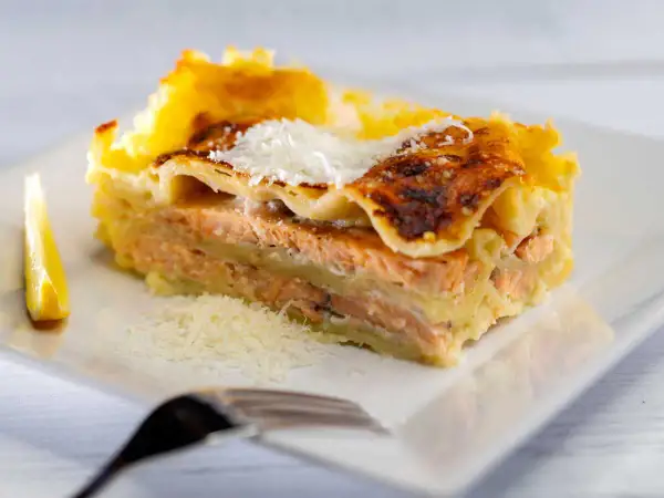 Une part généreuse de lasagne saumon, dorée au four, garnie de parmesan râpé et accompagnée d'un quartier de citron.