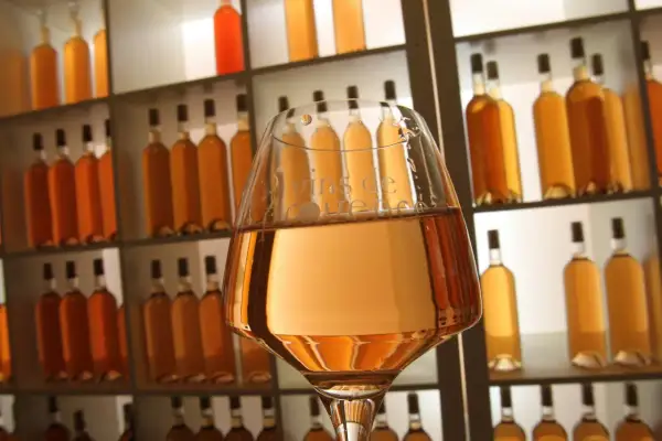 Verre de vin à la couleur rose très clair, devant une étagère remplie de bouteilles dorées.