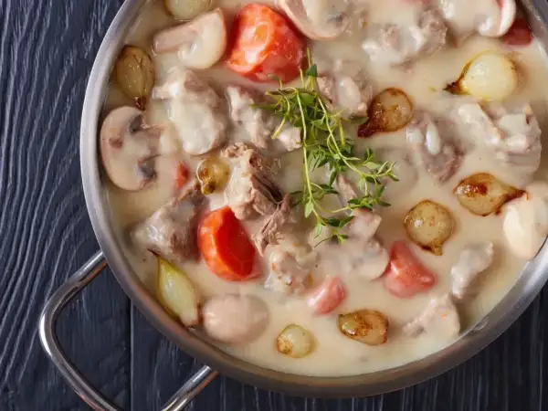 Un plat de blanquette de veau crémeuse avec des morceaux de viande tendre, des carottes, des champignons et des petits oignons, garni de thym frais.