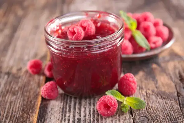 Un pot de confiture de framboise maison, garni de fruits frais et de feuilles de menthe, sur un fond de bois rustique.