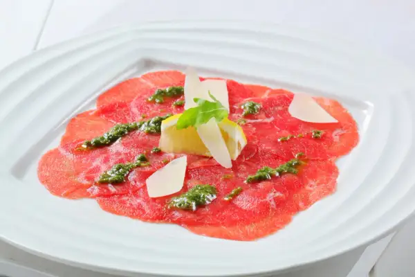 Carpaccio de bœuf finement tranché, arrosé d'une sauce verte et garni de copeaux de parmesan, de citron et d'une feuille de roquette.