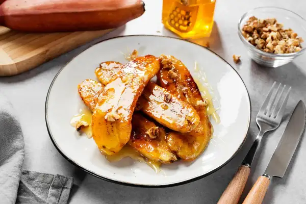 Bananes flambées caramélisées, nappées de miel et parsemées de noix, sur une assiette blanche. Un délice sucré.