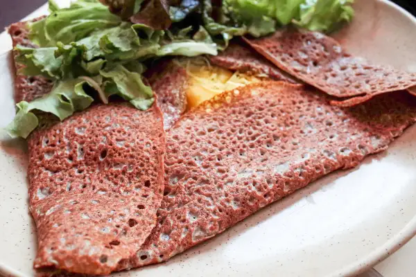 Une galette sarrasin pliée, garnie de fromage fondant et de salade verte fraîche, sur une assiette beige.