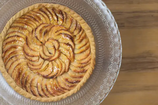 Une magnifique tarte aux pommes, aux pommes caramélisées disposées en spirale sur une pâte dorée, servie sur un plat en verre décoré.