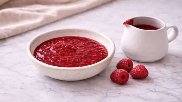 Un bol et un pichet remplis de coulis de framboise, avec trois framboises fraîches à côté.